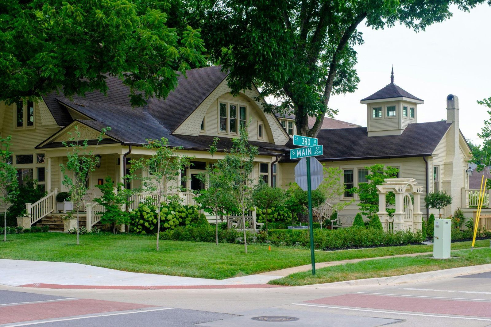 A cream-colored home on a corner lot beneath mature shade trees, with street signs reading SE 3rd St and S Main St